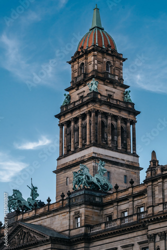 Close-up of the historic Wayne County Building in Detroit, Michigan, showcasing its ornate Beaux-Arts architecture, dome tower, and bronze quadriga statues against a blue sky.