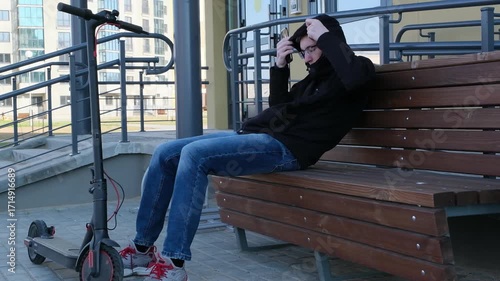 A young man rides an electric scooter along a paved path in an urban park with modern apartment buildings in the background.
