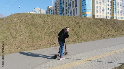 A young man rides an electric scooter along a paved path in an urban park with modern apartment buildings in the background.
