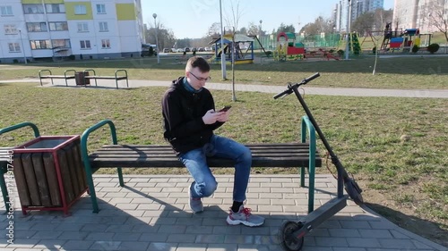 A young man rides an electric scooter along a paved path in an urban park with modern apartment buildings in the background.
