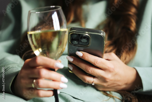 Woman holding wine glass and smartphone. Female hand with manicure multitasking. Relaxing indoors with technology. Drinking white wine while checking phone. Lifestyle with leisure and modern habits.