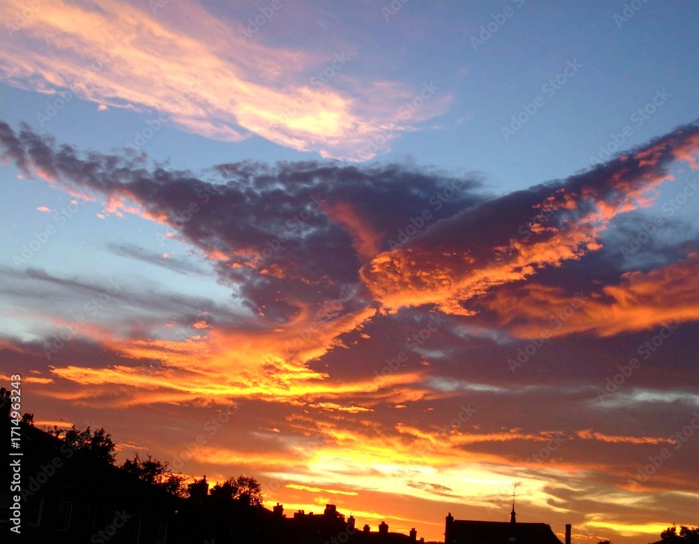 Fototapeta premium Fiery sunset clouds in molten bronze and indigo over silhouetted rooftops twilight