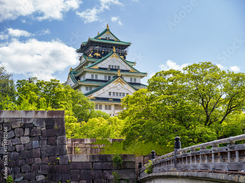 Osaka Castle surrounded by lush green trees and historic stone walls, under a bright blue sky with clouds in Osaka, Japan.