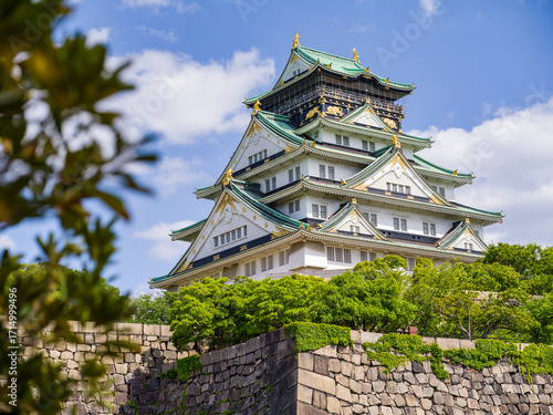 Osaka Castle viewed behind stone walls and green trees, with blue sky and clouds in Osaka, Japan.