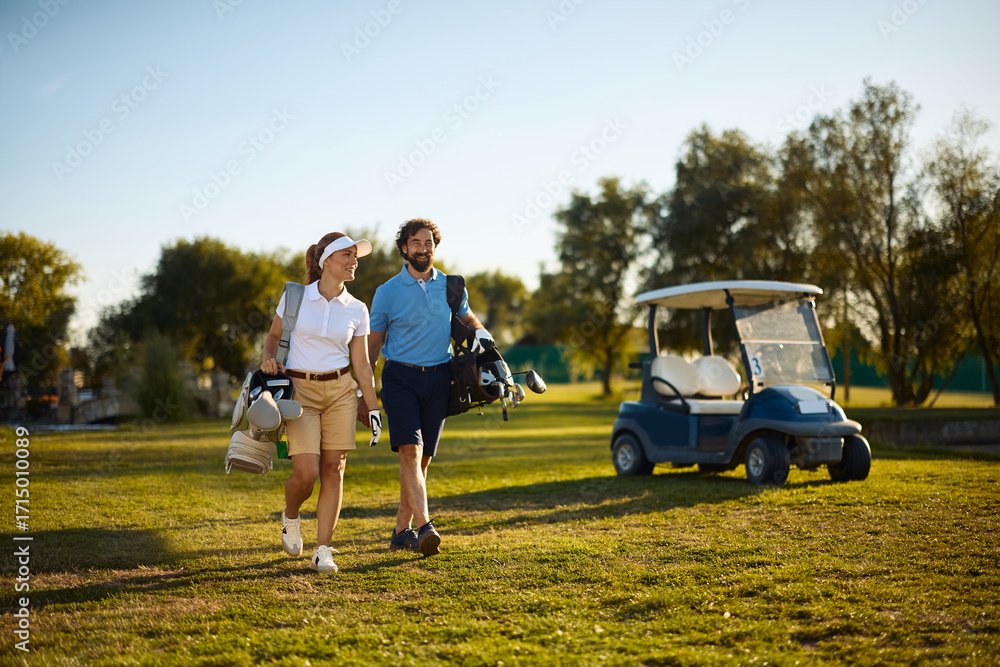 Fototapeta premium Happy couple of golfers carrying golf clubs in bags while walking on course.