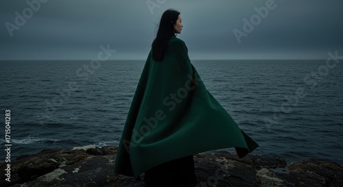 Woman in a billowing green cape viewed from the back stands on a rocky coast against a stormy ocean backdrop