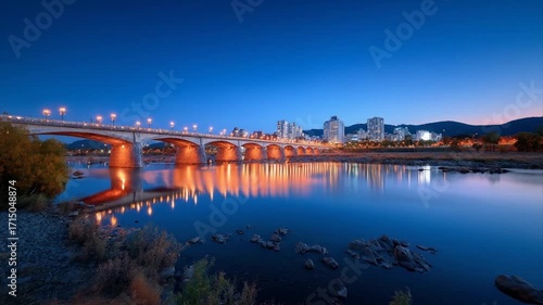 A tranquil cityscape at twilight, illuminated bridge reflecting on calm river waters.