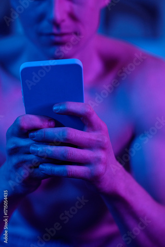 Man holding smartphone with both hands, focusing on device screen, upper body and face partially visible, illuminated by vibrant colored lighting, not looking at camera