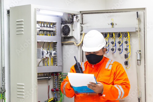 Electrician checking electrical panel safety while holding walkie-talkie and document, wearing PPE with hard hat and face mask ensuring protection against hazards and safety inspection protocols
