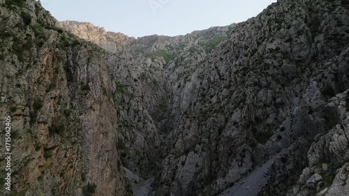 Morning drone flyover reveals a steep Uzbekistan gorge, limestone wall and a dark ravine with sparse alpine greenery. Soft dawn light touches the ridge and invites adventure in the Chimgan region