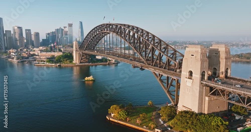Australia, Sydney: Aerial view of Sydney Harbour Bridge with cars driving on it, connecting the city skyline with the surrounding landscape, on a sunny morning with blue sky. Drone flight footage