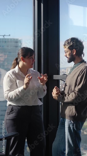 Two colleagues standing by a large office window having a casual conversation in natural light, representing workplace communication, connection and collaboration.