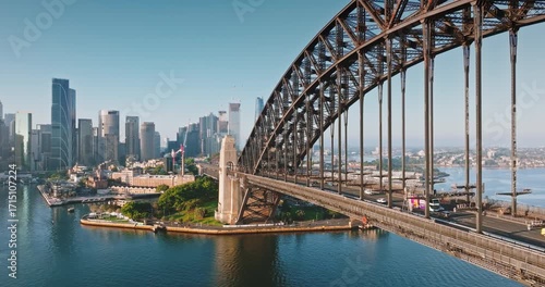 Australia, Sydney: Sydney Harbour Bridge in morning light. Cars driving traffic road connecting city skyline with surrounding urban landscape under bright blue sky. Aerial view drone flight footage