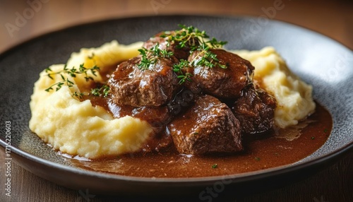 Plate of tender beef stew topped with rich brown gravy and fresh herbs served alongside creamy mashed potatoes on a dark ceramic plate