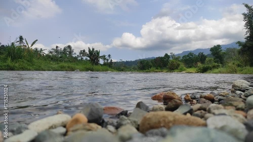 River Landscape with Stones: A serene river meanders through a picturesque landscape of lush greenery and rocky shores, under a sky dotted with fluffy clouds. Witness the harmony of nature.