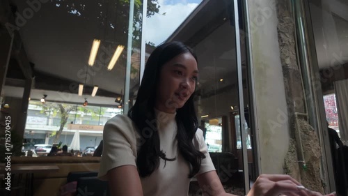 Beautiful Asian entrepreneur typing on her laptop while sitting at a wooden table in a cafe. The young woman is smiling and enjoying her work