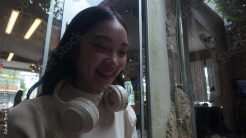 Young happy Asian woman with headphones using a mobile phone, texting, and smiling while sitting in a modern cafe near a large window