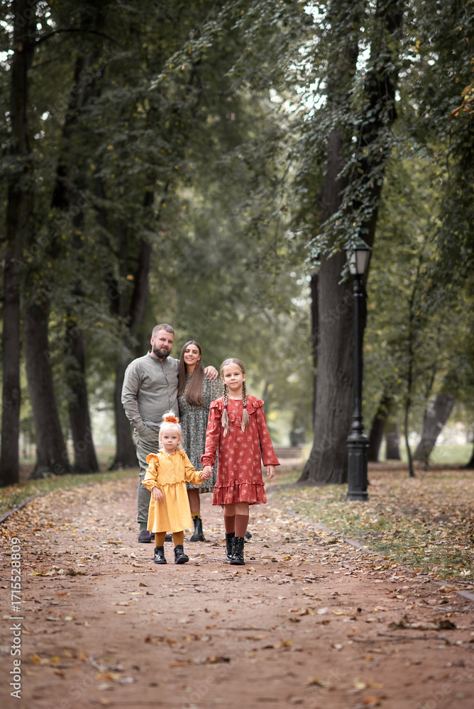 Fototapeta premium In an autumn park, two little girls are walking hand in hand on the road, while their parents stand embracing and watching them.
