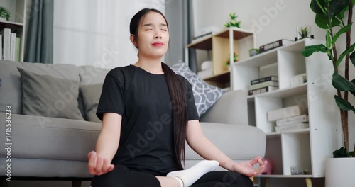 Handheld low angle view shot, Relaxed Asian woman practicing yoga by meditating on a yoga mat in living room at home. Self-care and mindfulness concept.
