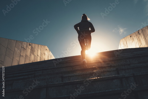 Low-angle shot of a woman running up long stadium stairs with sun flare behind her, dynamic composition symbolizing progress, perseverance, and determination in fitness and life.