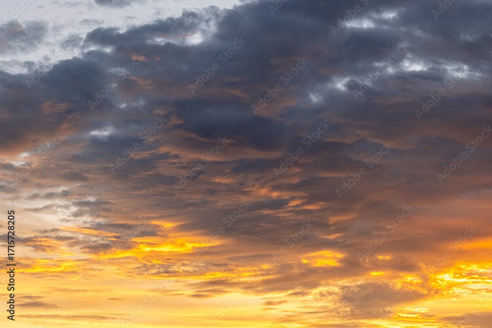 Obraz premium Dramatic sunset sky with golden and orange light illuminating dark clouds, creating a stunning natural background full of warmth, contrast, and atmospheric beauty.