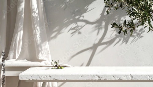 A minimalist  scene showcasing a simple white table and flowing white fabric draped over a white wall with olive branch shadows.