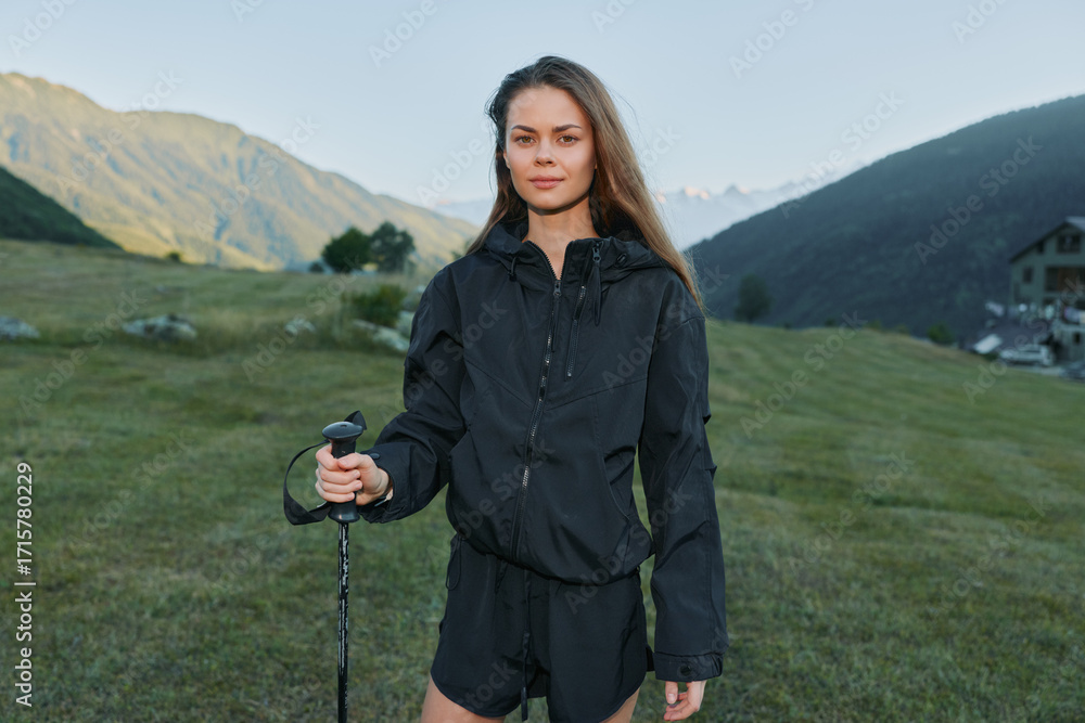 Fototapeta premium A confident young woman stands outdoors in a scenic mountain meadow, holding a trekking pole. She wears a dark jacket and shorts, looking ahead with calm determination, embodying adventure and
