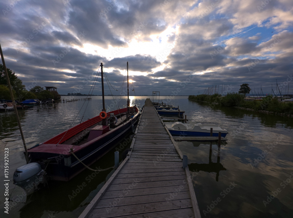 Obraz premium Wooden jetty with moored boats under a dramatic, cloudy sky at sunrise, Lake Dümmer in the morning in 49041 Damme, Germany