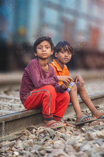 Two Boys Sitting on Railway Tracks Beside Moving Blue Freight Train in Slum Area