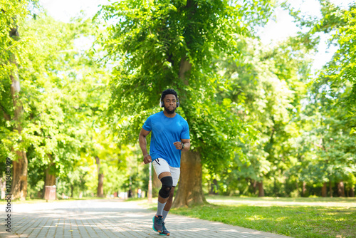 Schilderij op canvas Young african american man jogging in park with knee brace during workout