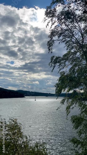 Static video of a sailboat moving on a lake with a twisted sail caused by wind. Forest and sky with clouds in background.