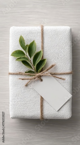 White gift wrapped in textured fabric with twine, green leaf sprig, and blank tag on wooden surface
