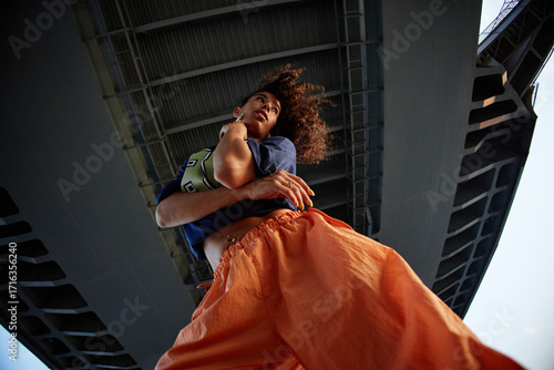 Bild auf Leinwand Young woman standing under bridge, looking away from camera, holding arm, curly