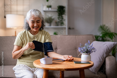 Asian elderly woman monitoring blood pressure at home for wellness senior health check self-care managing hypertension domestic living room medical device use