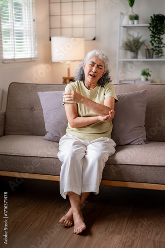 Older Asian woman experiencing arm pain on sofa at home aging joint issues chronic discomfort daily life challenges