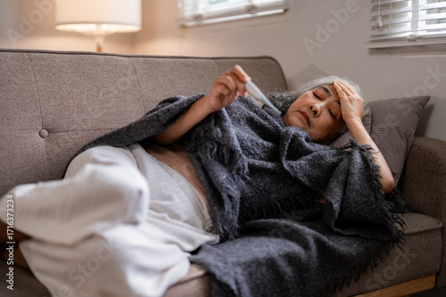 Canvastavla Older Asian woman with illness and fever resting on sofa measuring temperature a