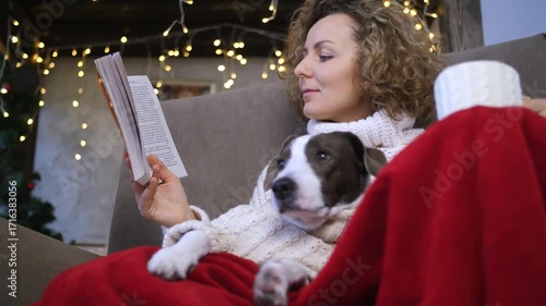 Christmas spirit enveloping spacious living room, woman reading book while nestling under soft red blanket, loyal dog resting nearby, warm golden lights illuminating peaceful holiday moment
