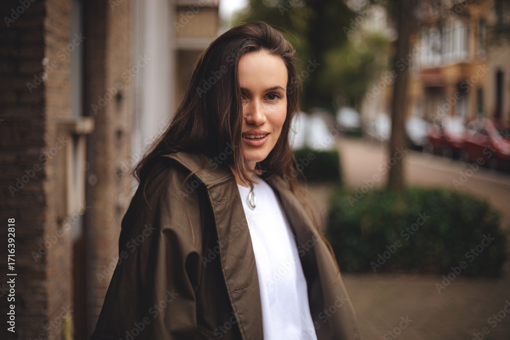 Fototapeta premium Woman wear trench coat. Portrait of young happy long hair brunette woman on city street looking at camera, laughing. Confident stylish girl outdoor. Proud and satisfied woman standing on street.