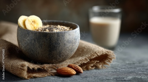 A bowl of healthy oatmeal topped with banana slices, chia seeds, and almonds, accompanied by a glass of milk on a textured surface.
