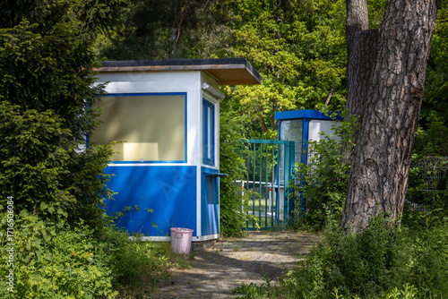 An Overgrown Abandoned Blue And White Ticket Booth With A Small Gate Situated Next To A Large Tree In A Forest