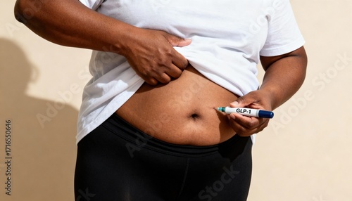 A middle-aged Black woman holds her shirt up, preparing to inject medication into her abdomen with a pen. She wears a white t-shirt and black leggings.