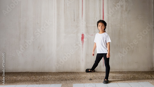 Portrait of cute Asian child in a simple white t-shirt and black leggings, standing. Background is a plain, old concrete wall with interesting red paint lines, creating a gritty, urban feel.