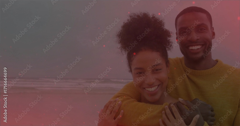 Naklejka premium Hugging couple wearing matching mustard sweaters at beach shoreline, ocean waves under cloudy sky