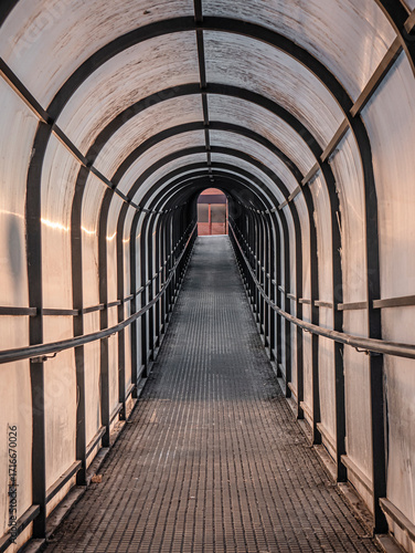 Pedestrian tunnel on an upward slope composed of a longitudinal arched metal structure of the same material and covered with a layer of translucent plastic through which the evening light enters