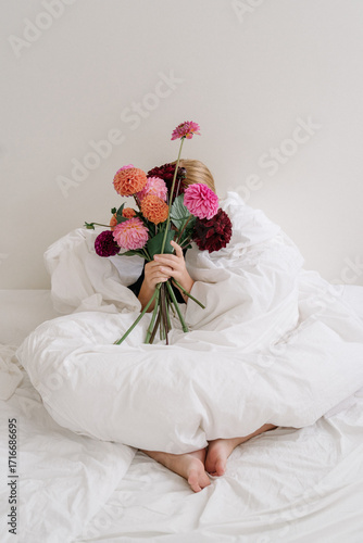 Girl sitting on bed wrapped in a blanket and holding a bouquet of burgundy, pink and orange dahlias. Minimalism, aesthetic