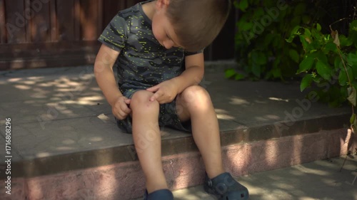 the child touches a scab on his knee with his finger while sitting on the porch on a hot summer day the camera slowly moves closer he is wearing a t shirt and shorts