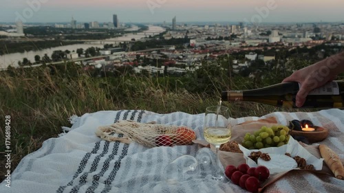 A cozy picnic on Vienna vineyards with wine being poured into a glass. Panoramic city view and sunset in the background create warm, romantic lifestyle and travel vibes.