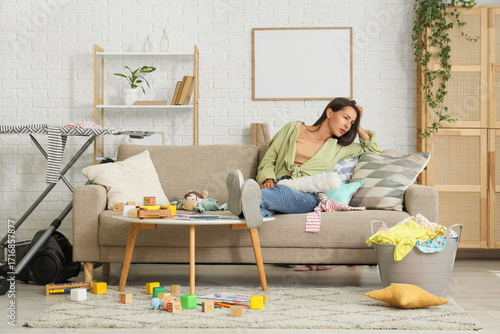 Quadro su tela Tired mother with scattered toys and basket of laundry lying on sofa in messy li