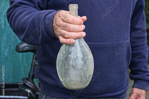 one man in blue clothes holding an old white empty dirty glass bottle in his hand on a summer street