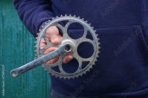 hand holds one detail of a sports bicycle made of gray iron bicycle sprockets with a knee for a pedal against the background of a green wall